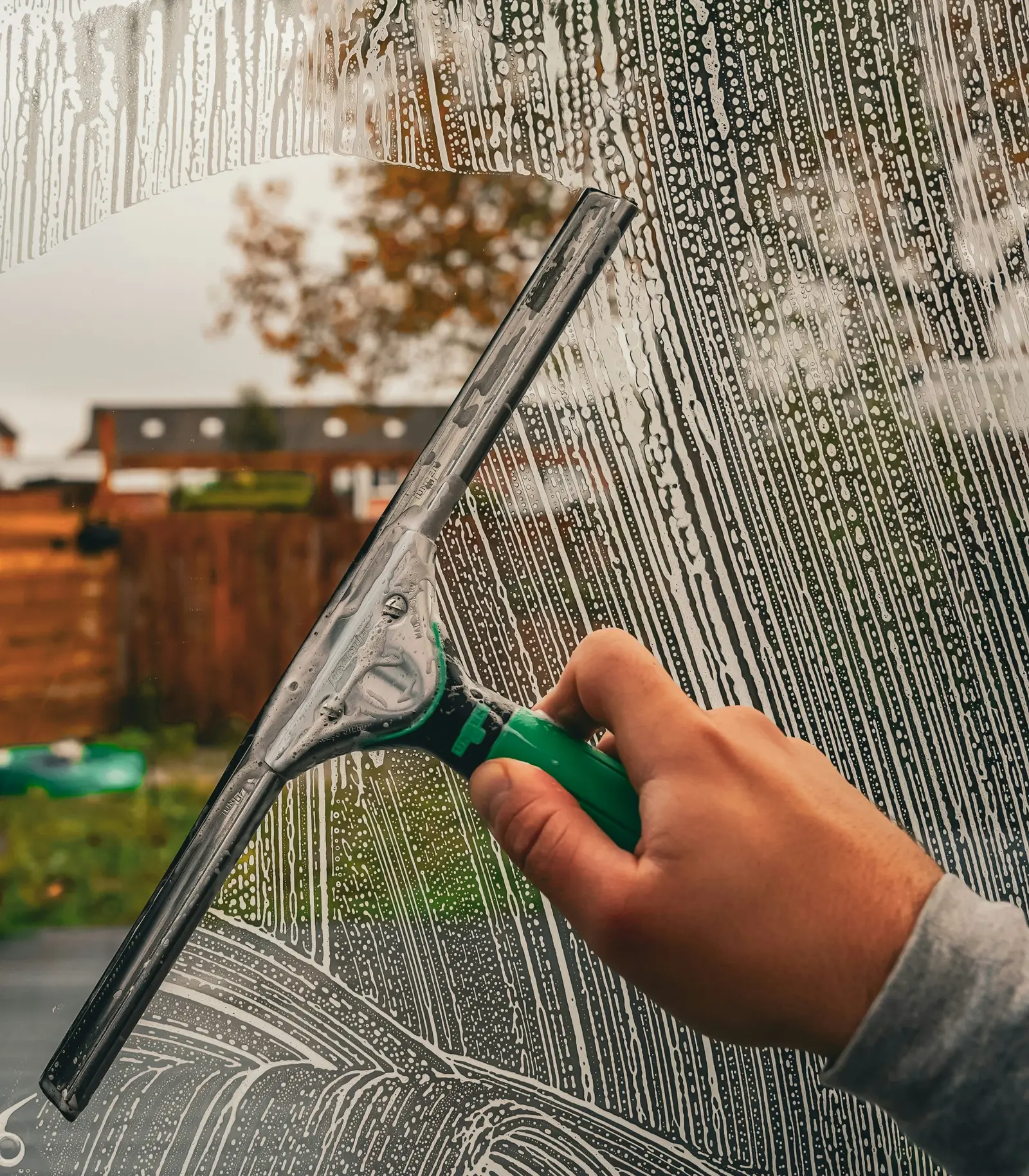 A hand running a squeegee down a soapy window during cleaning